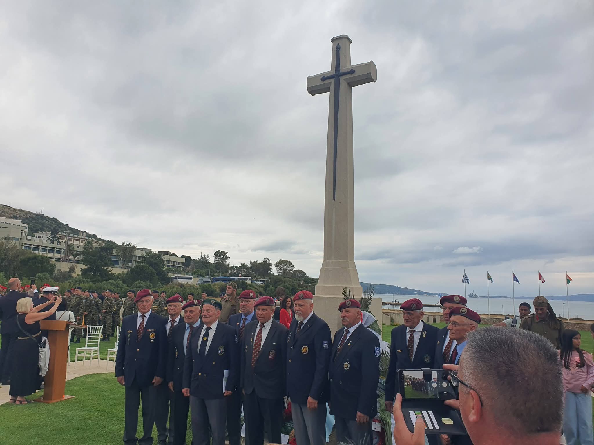Veterans in berets gathered before the Cross of Sacrifice at Souda cemetery, Battle of Crete commemoration
