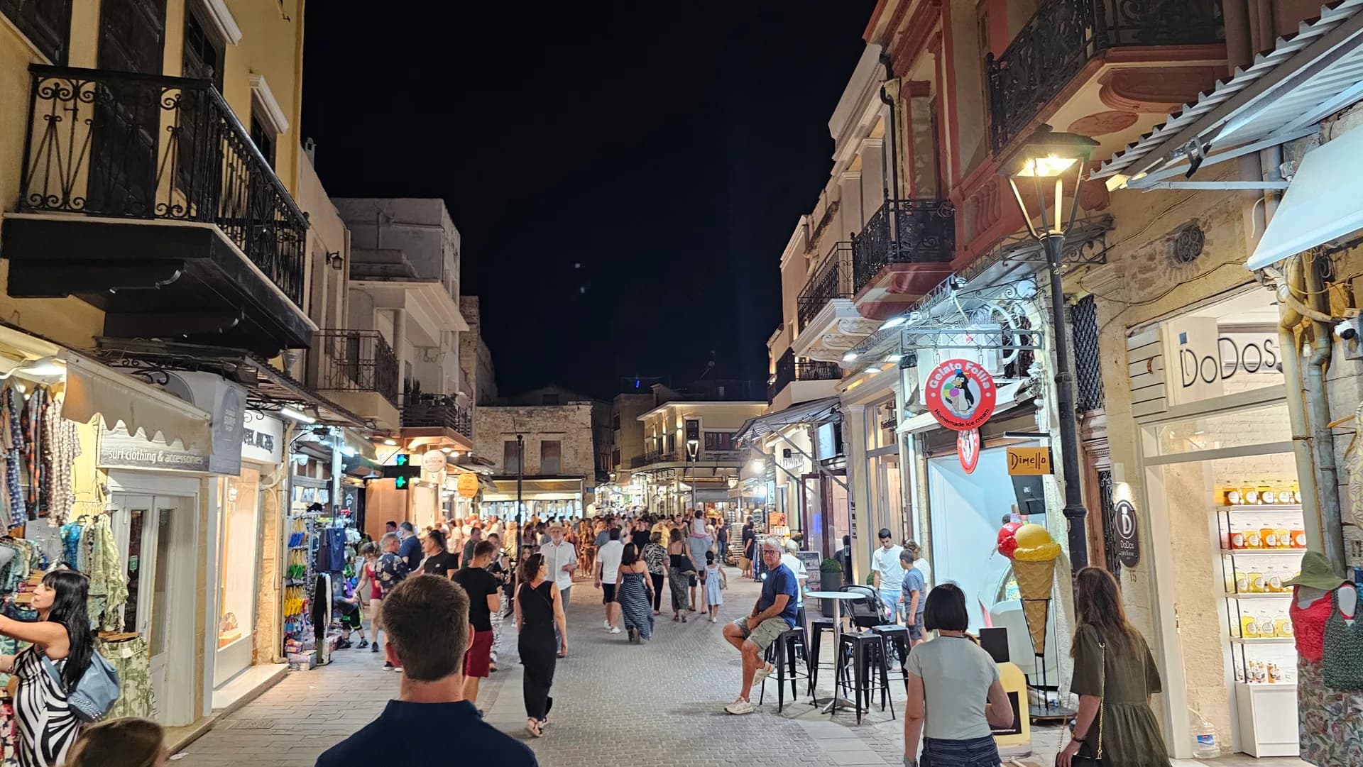 A shopping street in a Cretan old town at night, lit shopfronts and passers-by